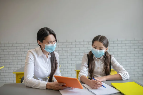 Lärare och skolflicka i masker sittande i klassrummet — Stockfoto