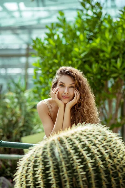 Pretty young woman standing near the cactus and looking thoughtful — Stock Photo, Image