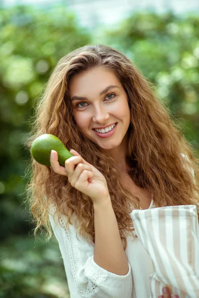 Beyaz gömlekli kadın avokado tutuyor ve gülümsüyor. — Stok fotoğraf