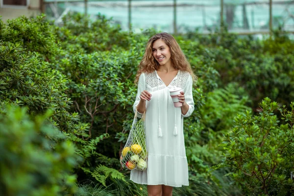 Mujer joven en vestido blanco con bolsas de verduras y una taza en las manos — Foto de Stock