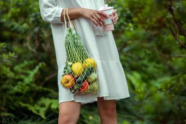 Close-up van vrouw in witte jurk met zakken van groenten en een kopje in handen — Stockfoto
