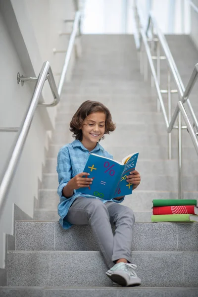 Mosolygó iskolás fiú ül a lépcsőn, matekkönyvet olvas — Stock Fotó