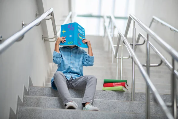 Schoolboy sitting on stairs, covering his face with math book — Stock Photo, Image