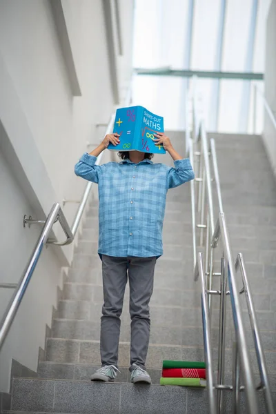 Schoolboy standing on stairs, closing his face with book — Stock Photo, Image