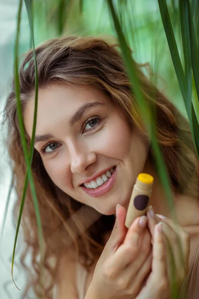 Mujer joven y bonita sosteniendo un palo de bálsamo labial y sonriendo —  Fotos de Stock