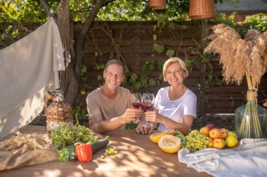 Orta yaşlı bir çift dışarıda masada oturmuş kırmızı şarapla kadeh kaldırıyor.