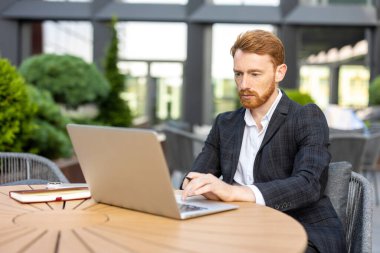 Kızıl saçlı iş adamı güneşli bir günde laptopla açık kafede çalışıyor.