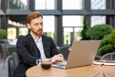 Kentsel bölgedeki sokak kafeteryasında dizüstü bilgisayarda yazan kızıl sakallı adam.