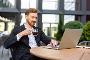 Sakallı, kızıl saçlı girişimci teras masasında kahve molası veriyor.