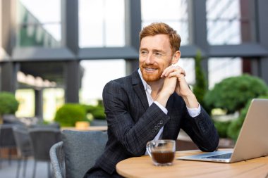 Kızıl saçlı adam dışarıda dizüstü bilgisayarın başında otururken gözlerini kaçırıyor.