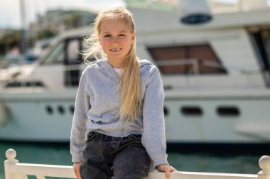 Sarışın kız rıhtımın kenarında oturuyor. Yatların yanında. Gün ışığında gülümsüyor.