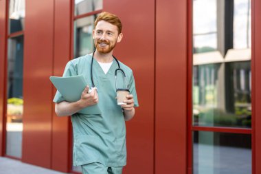 Ginger man doctor with beard walking in scrubs holding coffee and folder