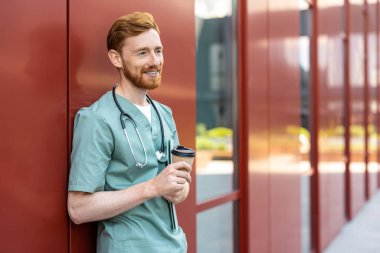 Ginger man doctor in scrubs drinking takeaway coffee near red hospital wall