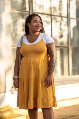 Smiling female with tattoo on arm posing by urban building in mustard dress enjoying sunny day