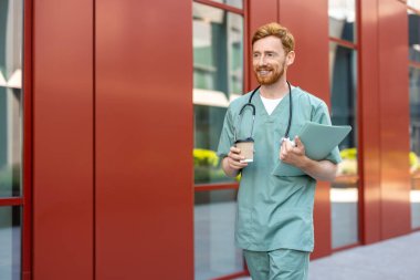 Bearded male medic carrying coffee cup and documents near hospital building