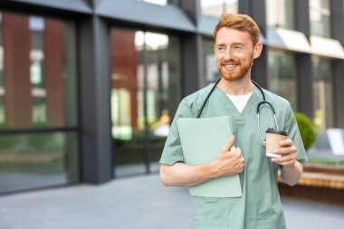Red-haired man physician in scrubs standing near hospital with cup and folder