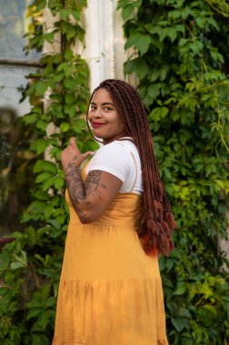 Smiling woman with braided hair in yellow dress standing outdoors near green plants