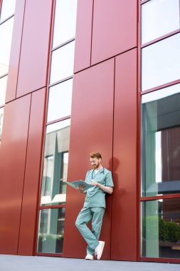 Bearded man in scrubs standing against red wall reading documents