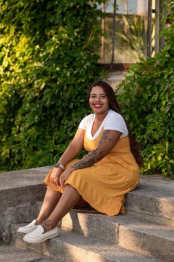 Smiling woman in yellow dress with braids sitting on outdoor stairs near building enjoying summer day