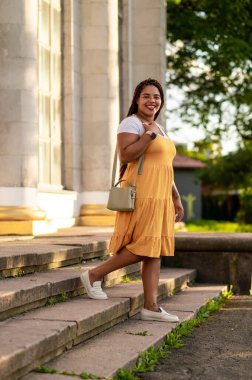 Smiling female with tattoo on arm climbing stairs in mustard dress enjoying sunny outdoor