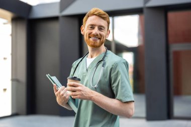 Caucasian man in scrubs carrying coffee and paper folder in urban setting