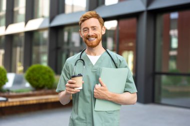 Smiling man in scrubs holding takeaway coffee and medical folder near clinic
