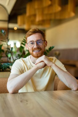 Smiling redhead resting arms on table in cozy cafe interior waiting his order