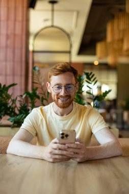 Smiling redhead man with braces looking at smartphone in cafe