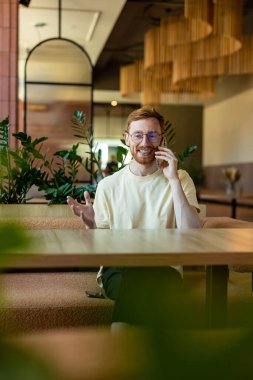 Smiling ginger man making phone call while seated at wooden table in cafe