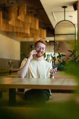 Red-haired man in glasses communicating on phone in cozy cafe