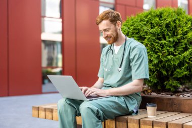 Smiling man doctor in scrubs sitting outdoors with laptop on lap and cup on bench