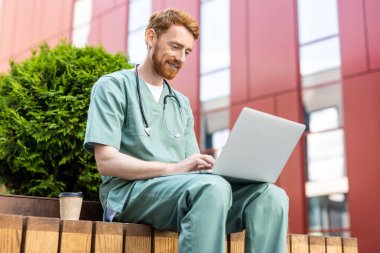 Red-haired man in medical uniform using laptop while drinking coffee outside