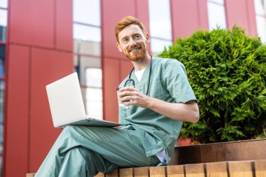 Smiling man in scrubs holding takeaway drink while using laptop near building