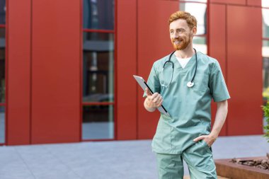 Red-haired man in medical uniform standing near hospital with laptop in hands