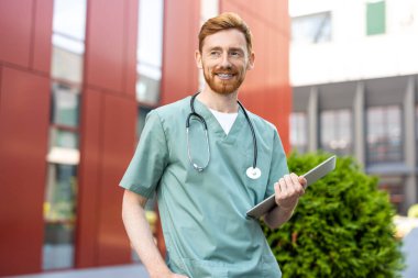 Smiling man in green scrubs carrying laptop in hands near hospital wall