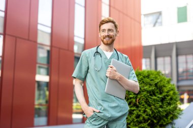 Ginger bearded man in scrubs holding closed laptop in front of building