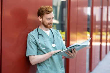 Man doctor in scrubs standing by wall reading from open medical folder