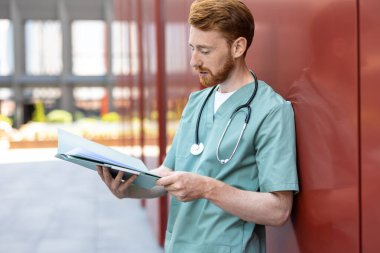 Bearded man in green uniform leaning on wall with open patient records