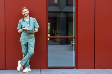 Ginger bearded man in scrubs standing against wall reading documents