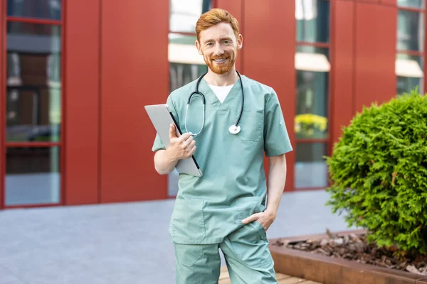 Smiling man doctor in scrubs standing outdoors holding closed laptop in hands