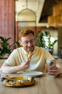 Bearded man with ginger hair in casual shirt using mobile while dining in cafe