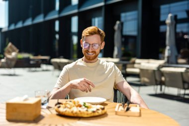 Handsome young ginger man wearing casual T-shirt and glasses eating pizza in outdoor cafe