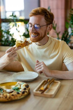 Ginger man smiling while enjoying pizza in bright modern dining place