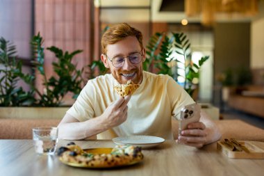Ginger bearded young adult in glasses holding phone and enjoying pizza indoors