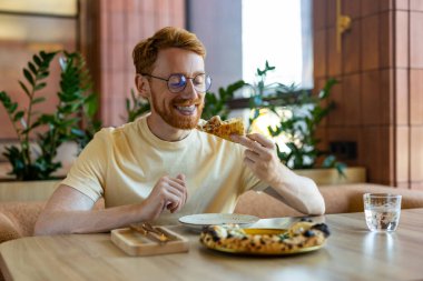 Bearded red-haired man in yellow shirt sitting at cafe table with pizza