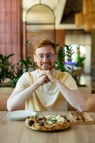 Ginger bearded man in glasses enjoying pizza while dining in cafe