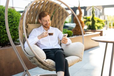 Gülümseyen kızıl saçlı adam kahve fincanı tutuyor ve rahat bir şekilde dışarıda otururken akıllı telefonu kontrol ediyor.