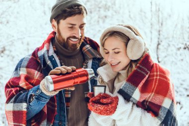 Kış ormanında sıcak çay. Gülümseyen adam, kız arkadaşının fincanına sıcak çay dolduruyor ve ikisi de keyifli görünüyor.