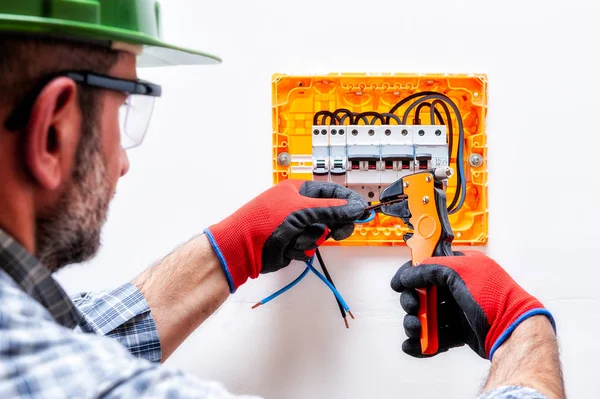 Electrician technician with helmet, goggles and gloves protected hands, works with wire stripper in a residential electrical panel.