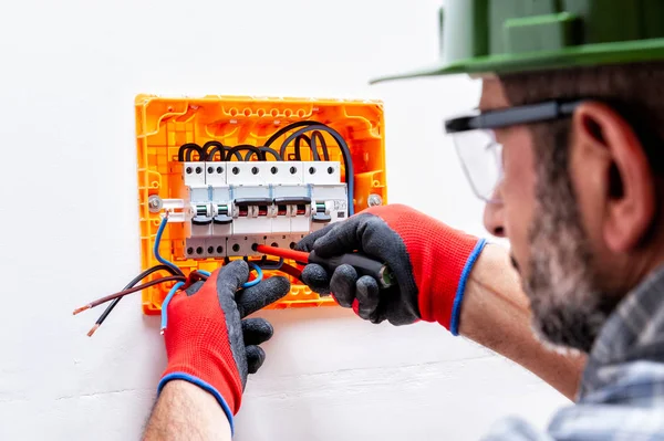 Electrician technician with helmet, goggles and hands protected by gloves with screwdriver fastens the cable in a residential electrical panel.
